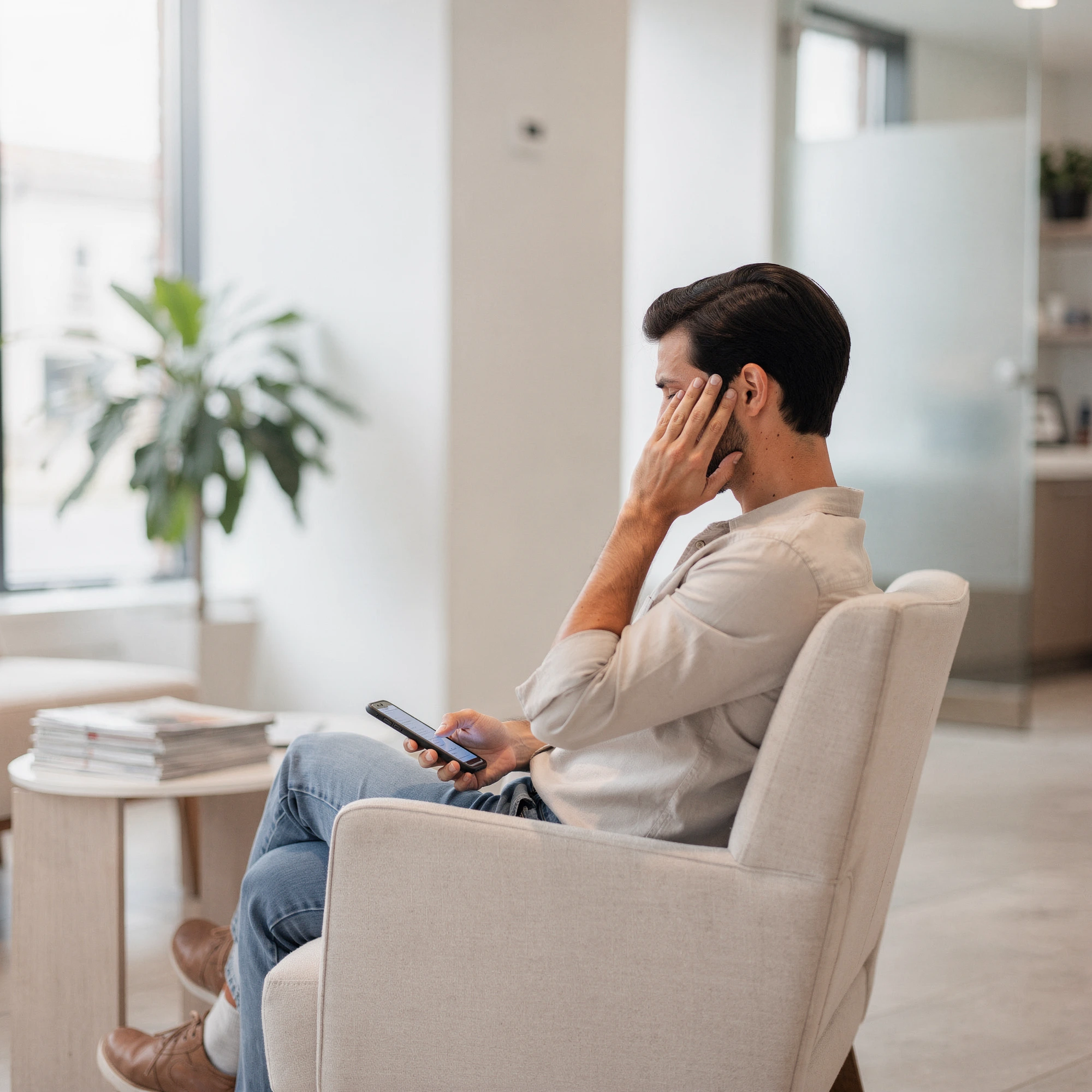 Man in dental waiting room holding phone and touching his cheek in discomfort